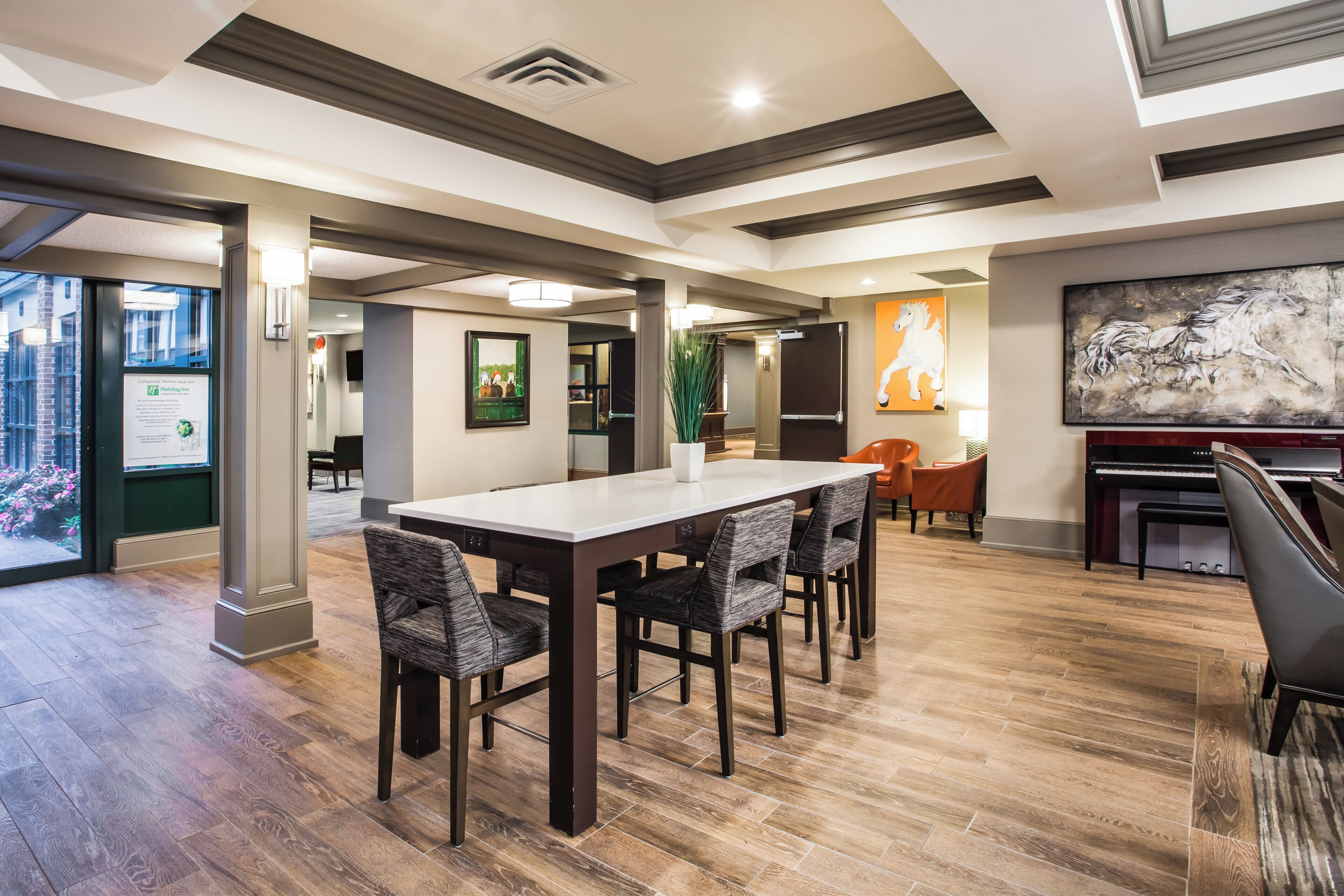 A modern hotel lobby with a dining area featuring a long table and chairs, a large painting on the wall, and a view of the outside through a glass door.