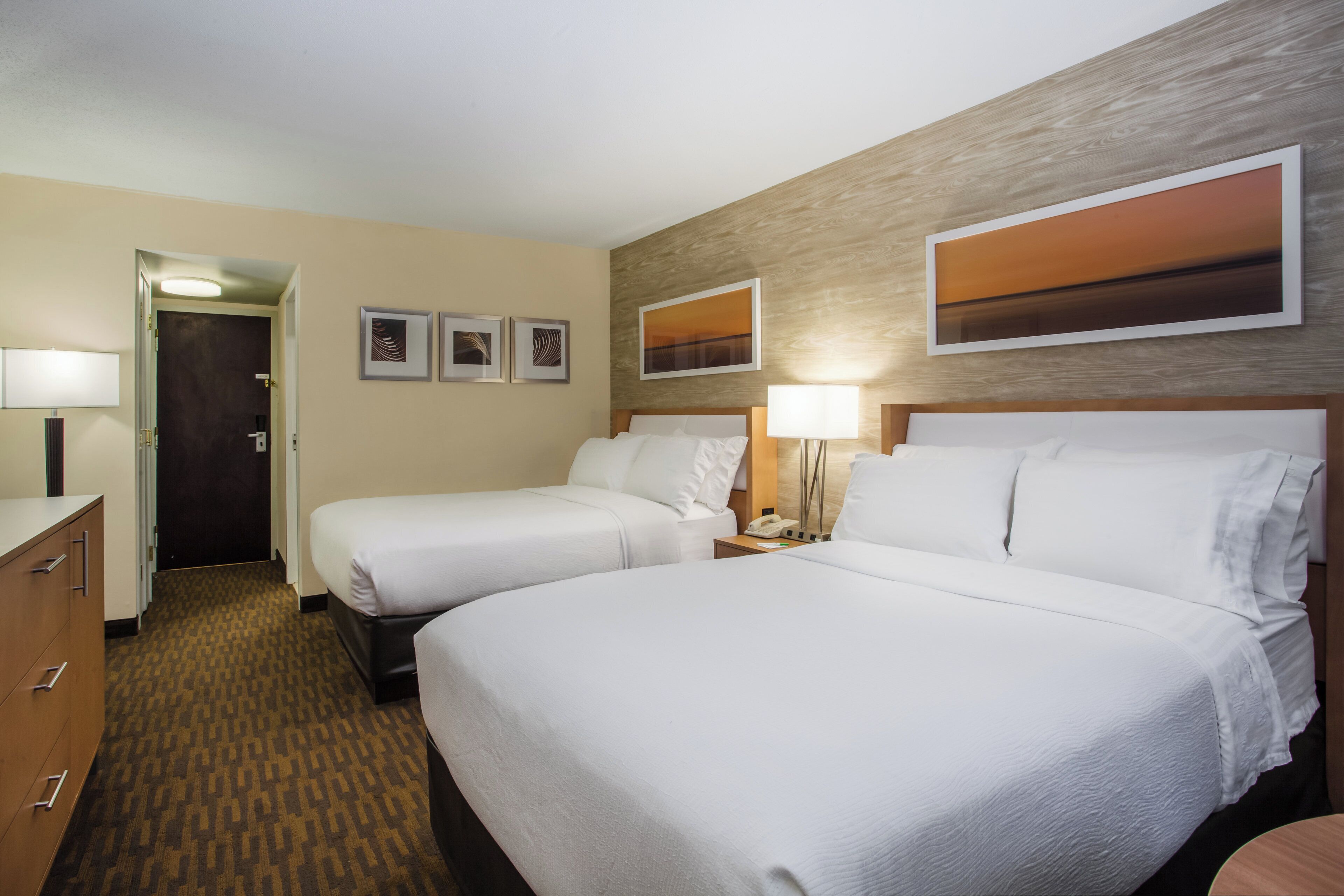 A hotel room with two beds, a dresser, a desk, and a door leading to a hallway.