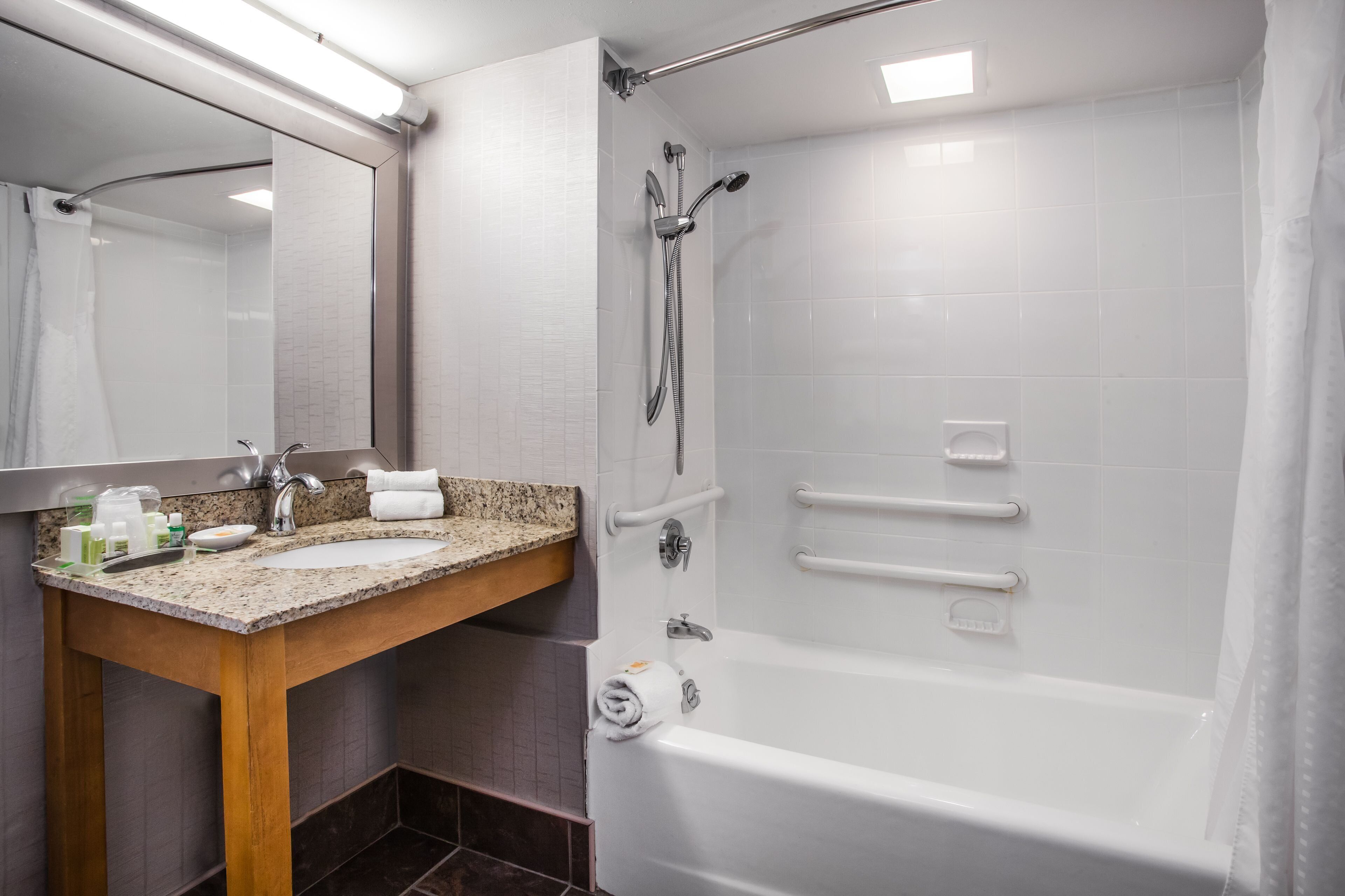 A modern bathroom with a granite countertop sink, a large mirror, a bathtub, and a shower with grab bars.