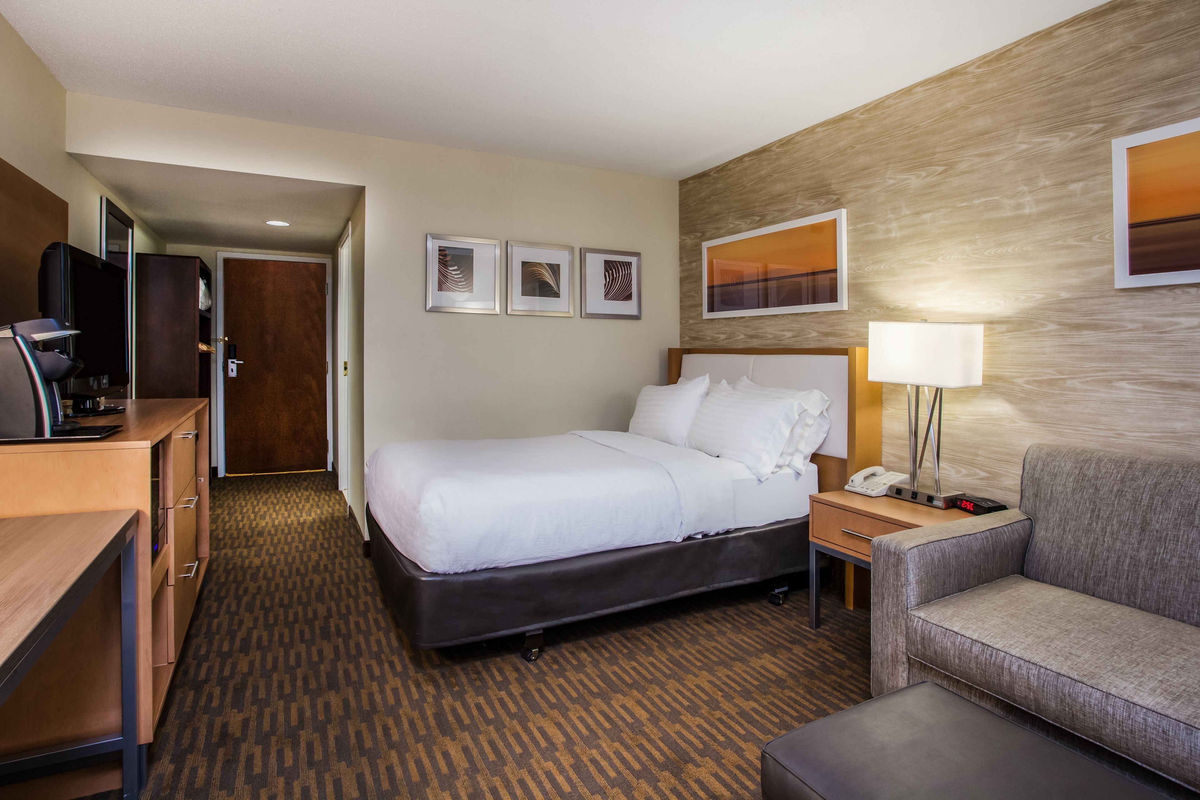 A hotel room with a bed, a desk, a chair, a lamp, and artwork on the walls.