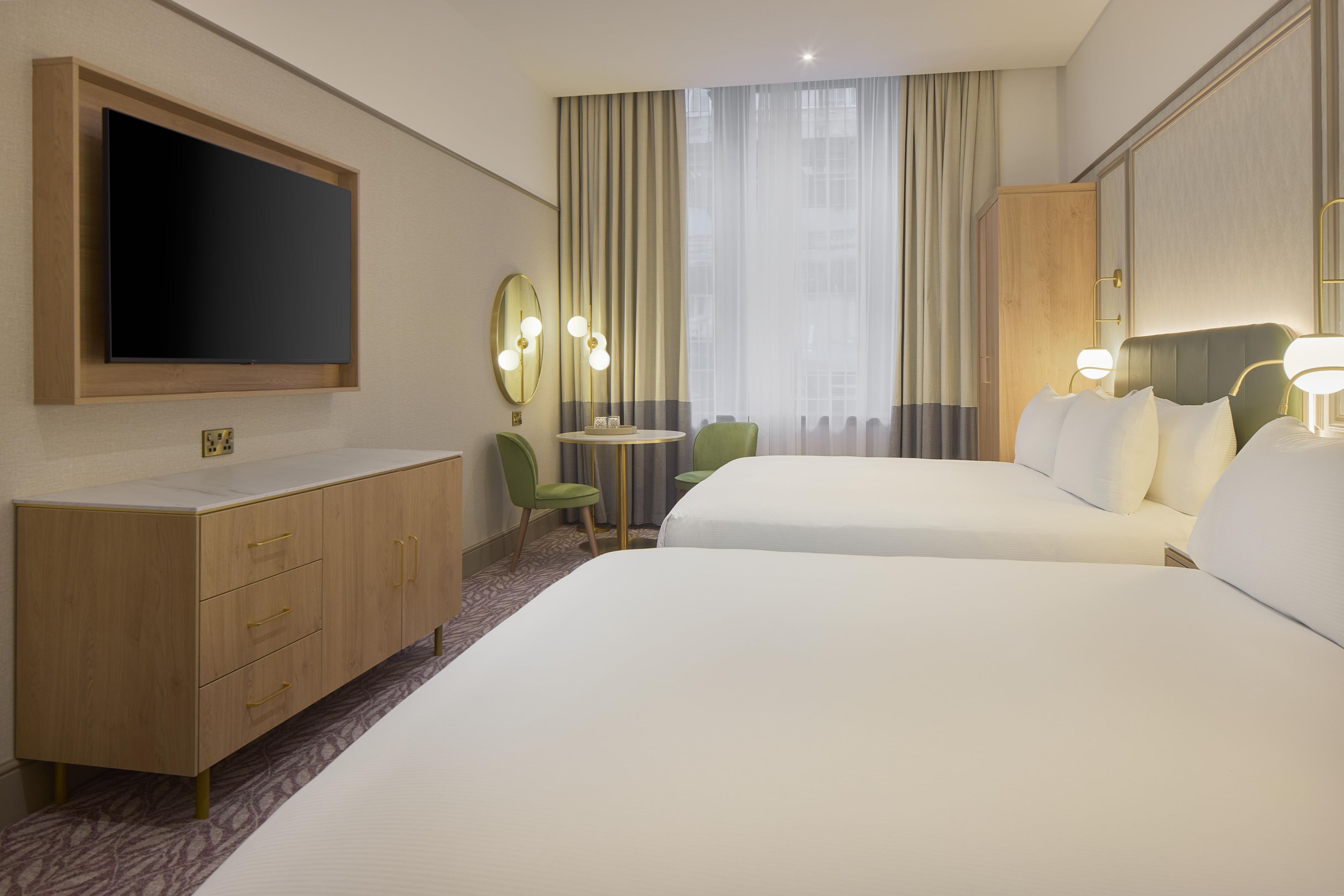 A hotel room with a TV, a desk with a chair, two beds, and a large window with curtains.