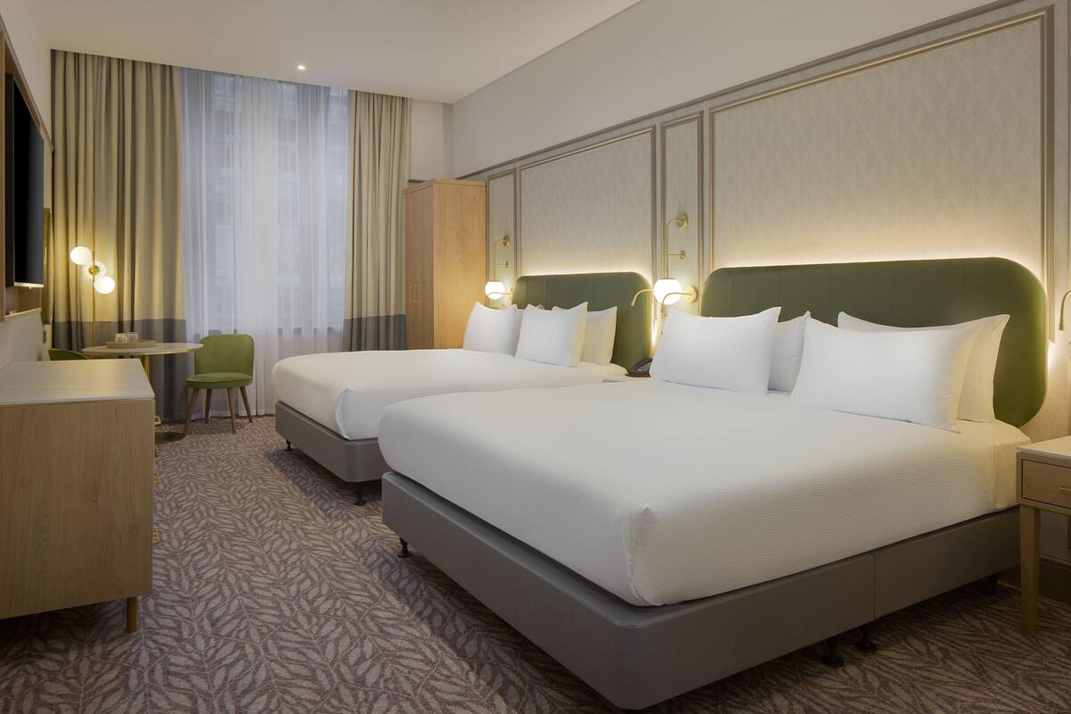 A hotel room with two beds, a desk, a chair, and a large window with curtains.