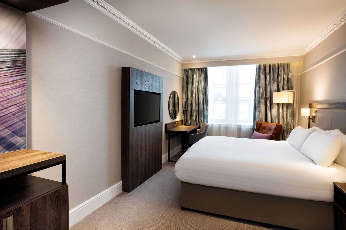 A hotel room with a large bed, a desk, a TV, and a window with curtains.