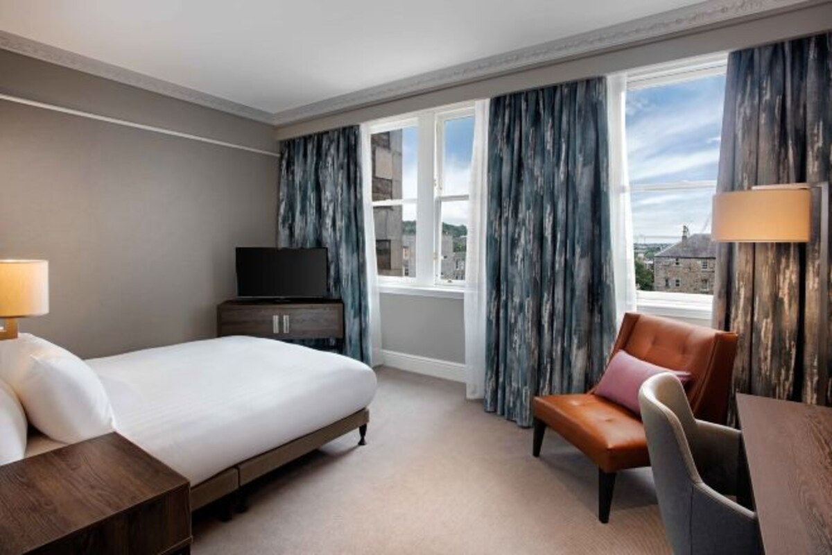 A hotel room with a bed, a desk, a chair, a TV, and a window with curtains.
