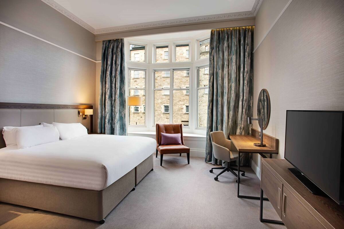 A hotel room with a large bed, a desk with a chair, a television, and a window with curtains.