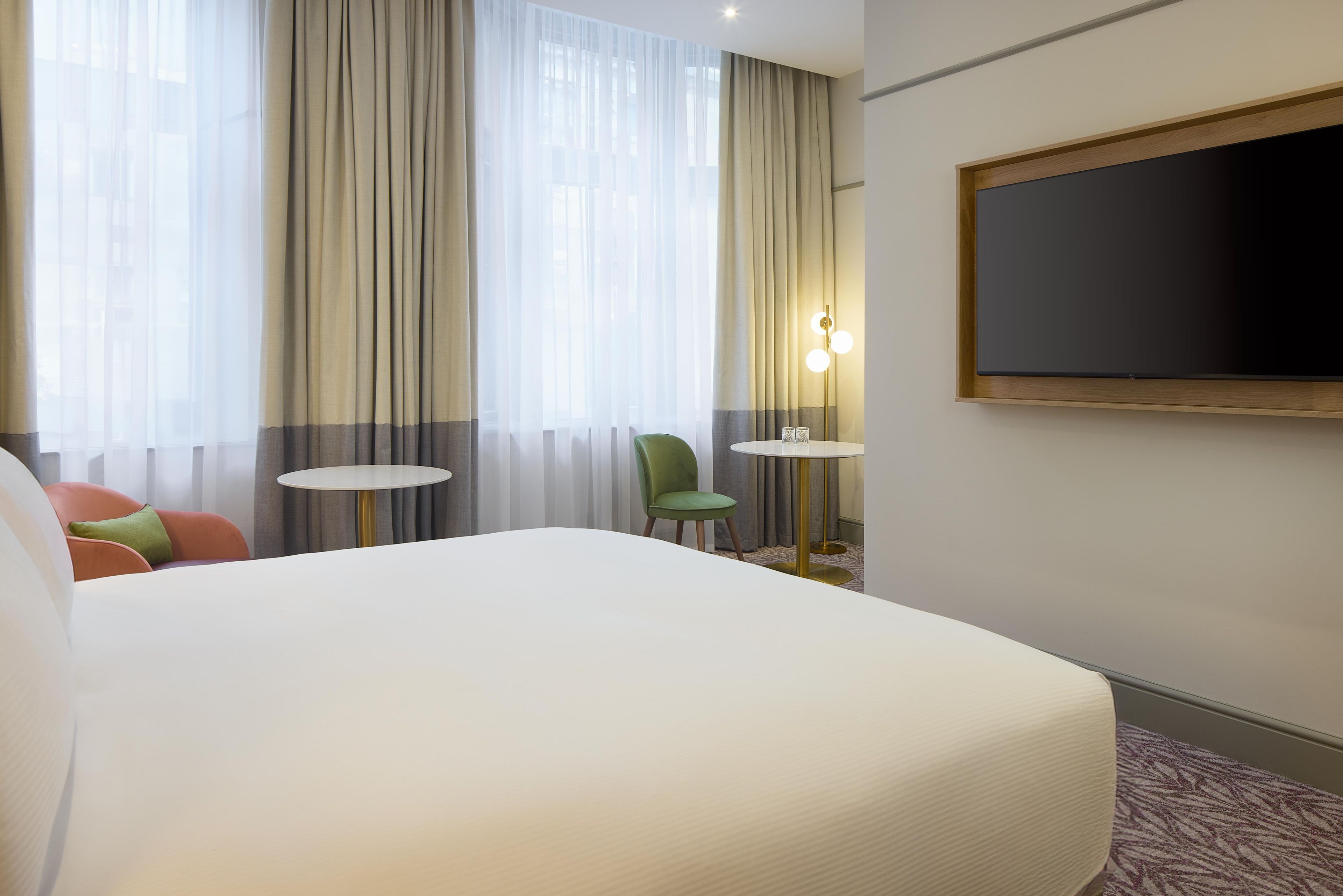 A hotel room with a large bed, a small round table, a green chair, and a TV mounted on the wall.
