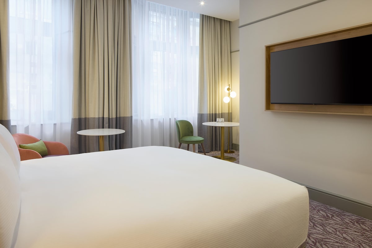 A hotel room with a large bed, a small round table, a green chair, and a TV mounted on the wall.