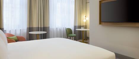 A hotel room with a large bed, a small round table, a green chair, and a TV mounted on the wall.
