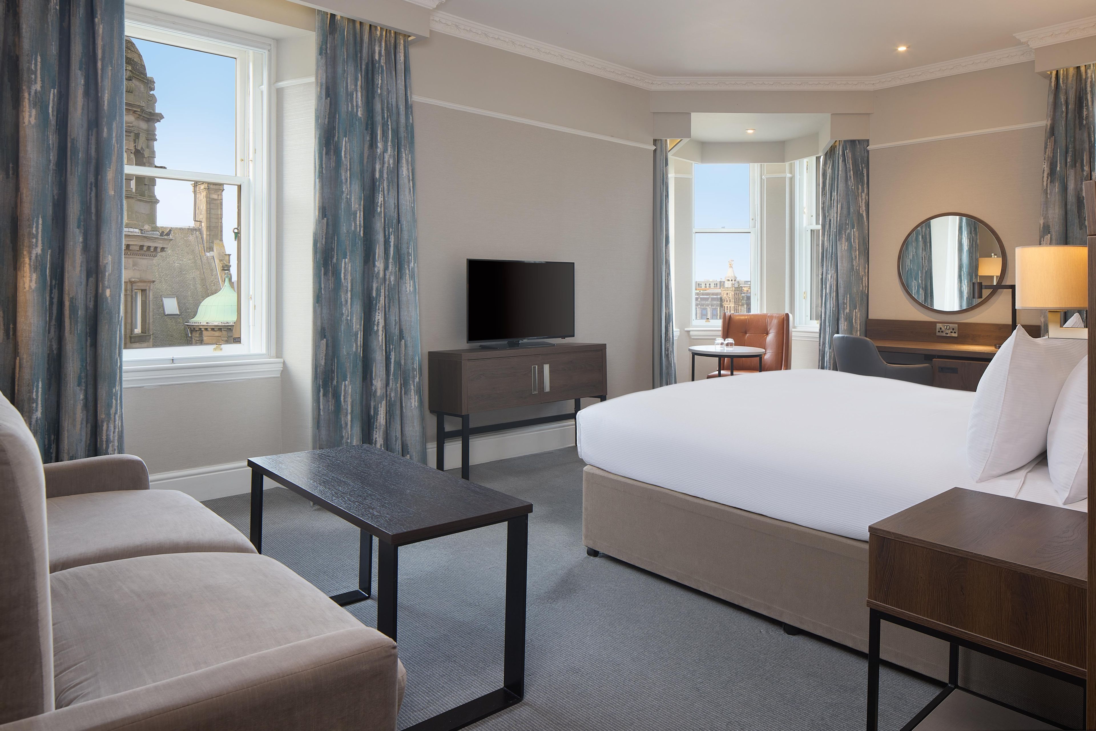 A hotel room with a bed, a television, a desk, a chair, and a view of the city.