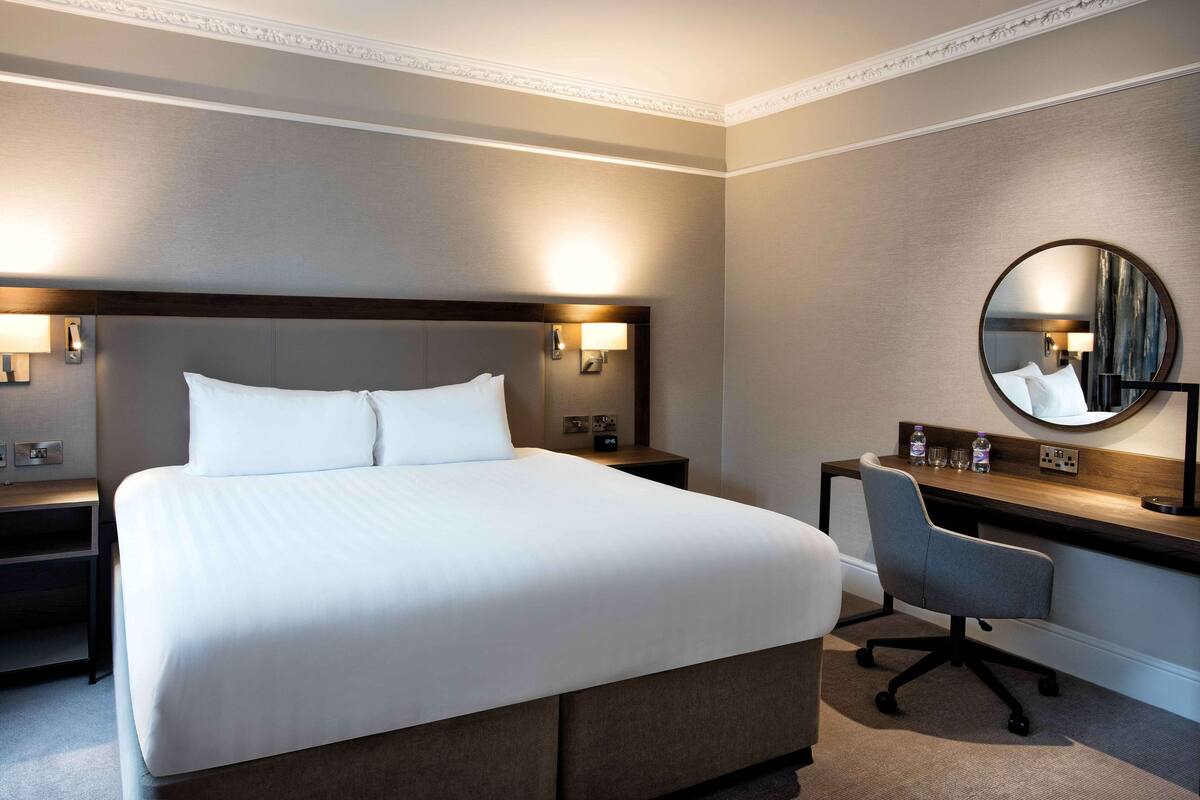 A hotel room with a large bed, a desk, a chair, a round mirror, and two bedside tables with lamps.
