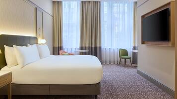 Quarto de hotel com uma cama grande, televisão, uma mesinha e uma cadeira.