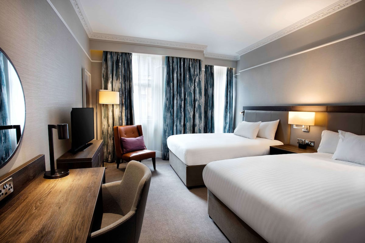 A hotel room with two beds, a desk with a TV, a chair, and a large window with curtains.