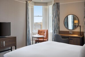 A hotel room with a bed, a desk, a chair, a television, a round mirror, and a view of a cityscape through a window. - Hilton Edinburgh Carlton (Edinburgh)