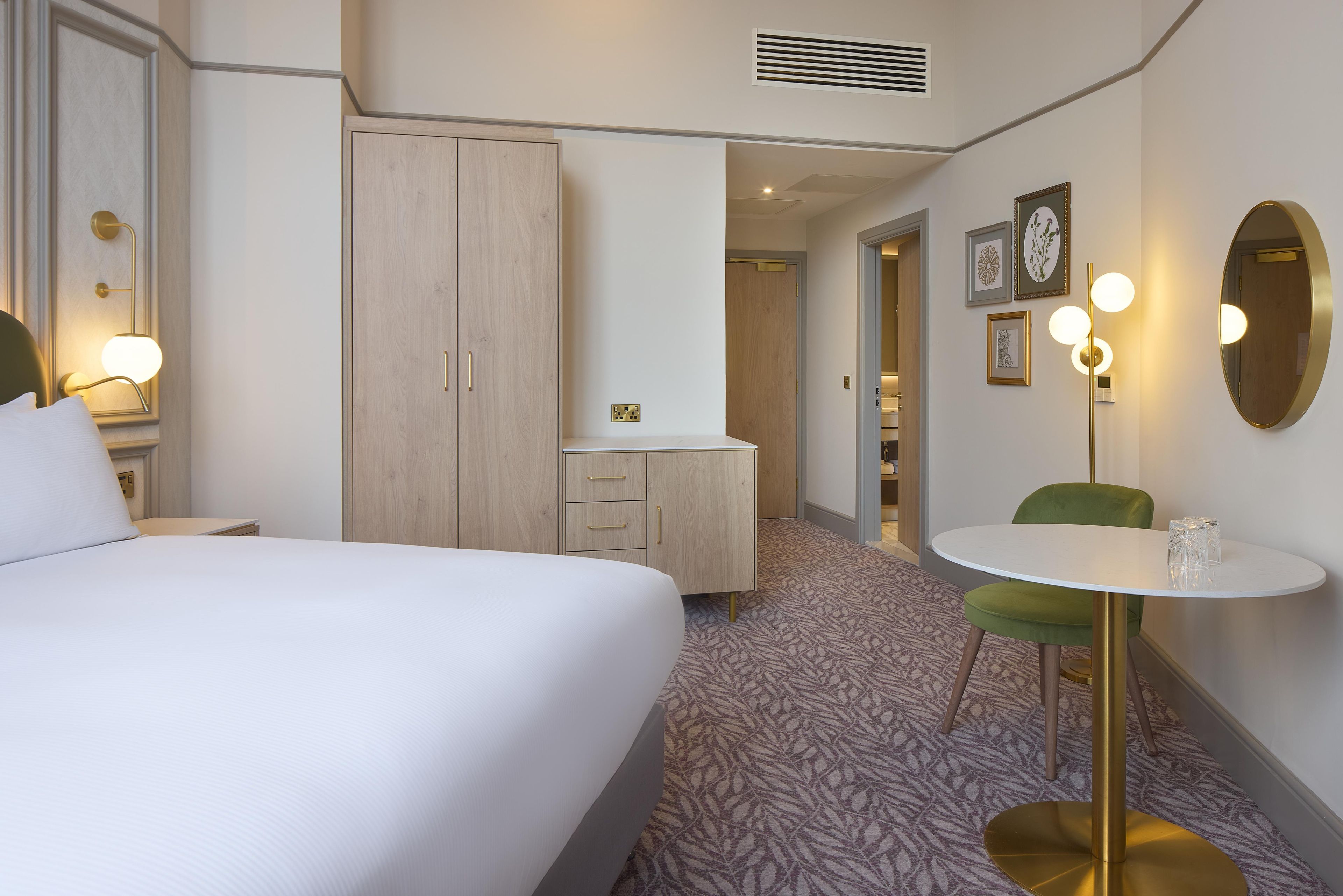 Ein Hotelzimmer mit einem Bett, einem Schreibtisch mit Schubladen, einem grünen Sessel, einem runden Tisch und einem Spiegel.