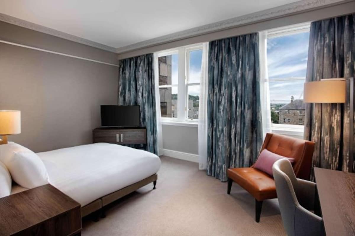 A hotel room with a bed, a desk, a chair, a television, and a window with curtains.
