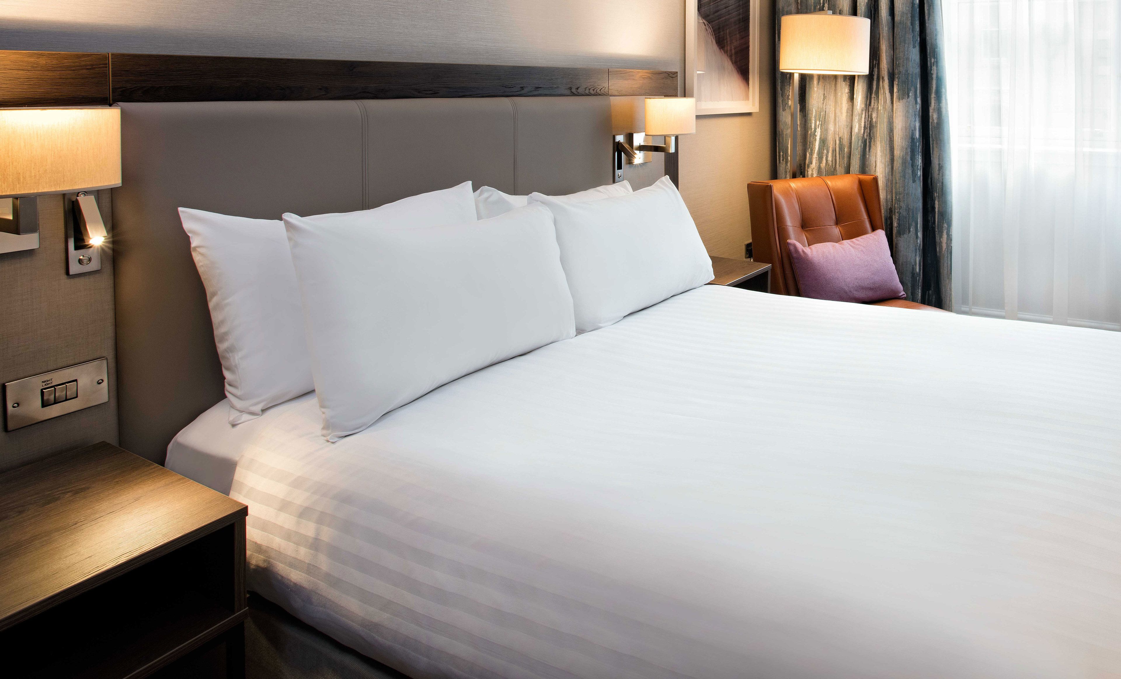 A hotel room with a large bed, a night switch, a lamp, and a chair.