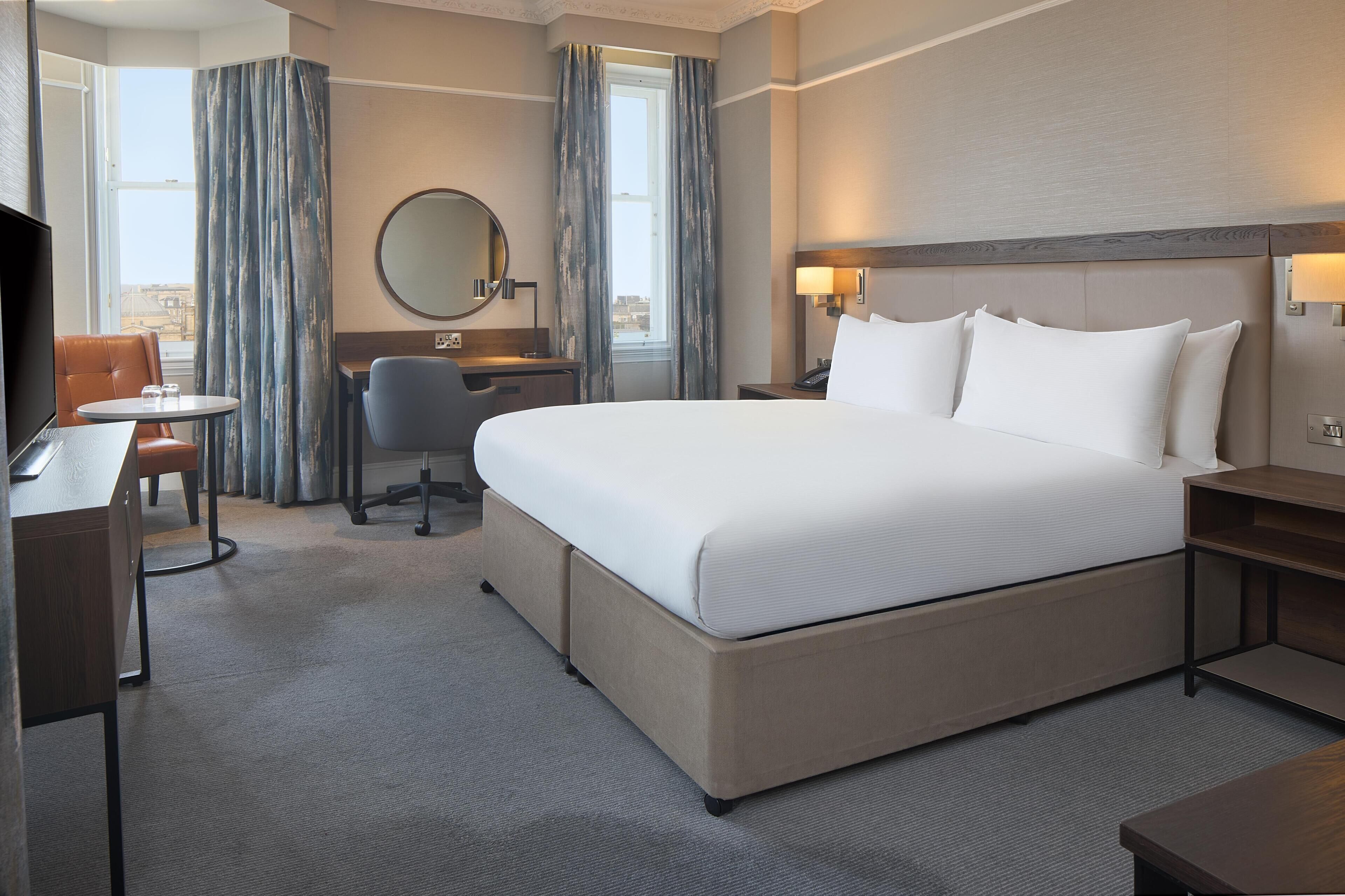 A hotel room with a large bed, a desk, a chair, a round mirror, and a view of the city through the window.