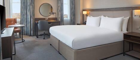 A hotel room with a large bed, a desk, a chair, a round mirror, and a view of the city through the window.