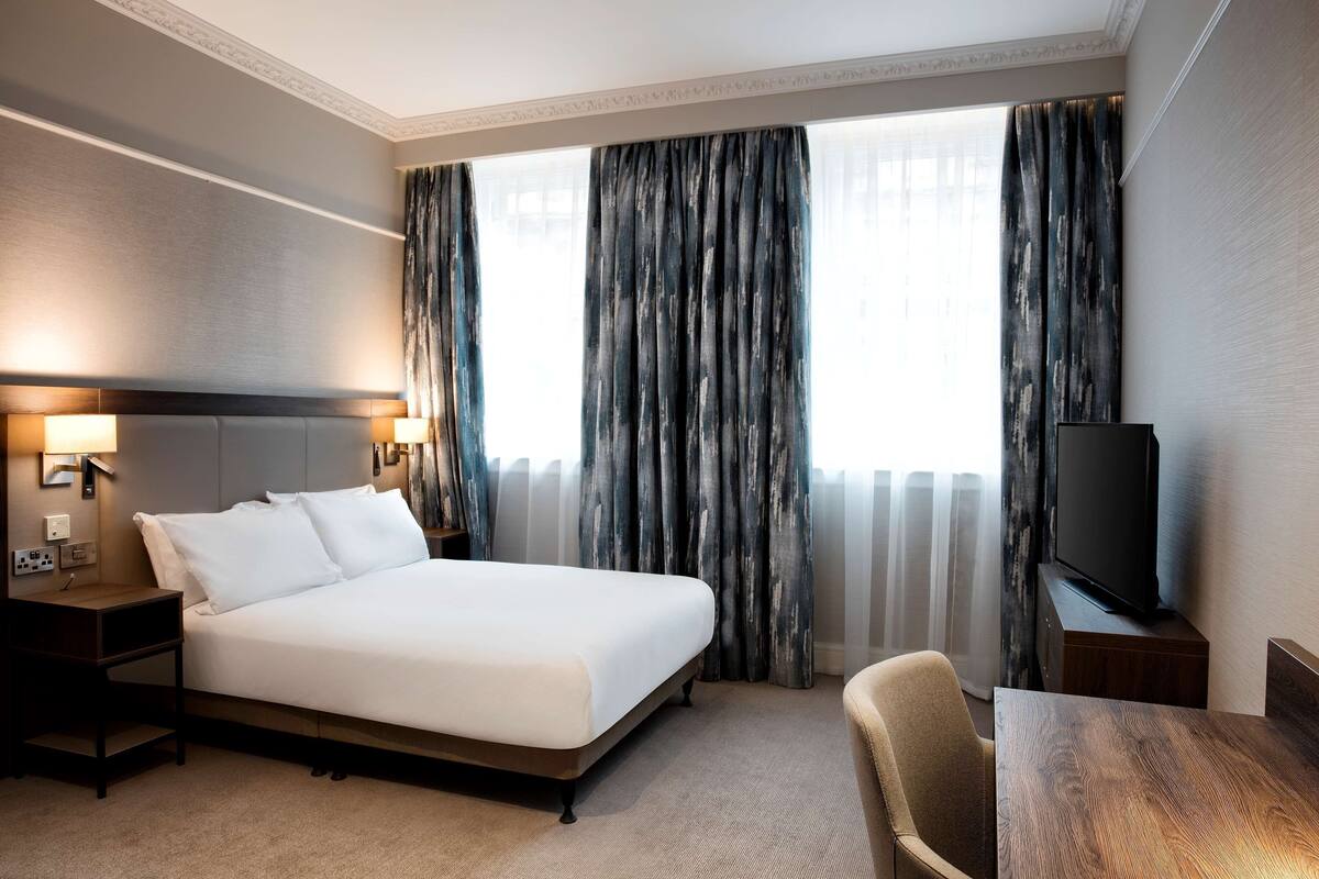 A hotel room with a large bed, a TV on a stand, a desk, and a chair.