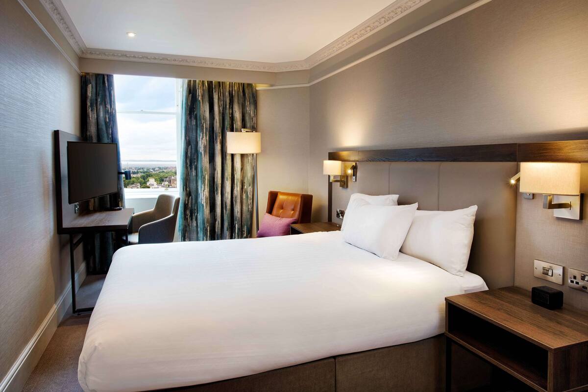 A hotel room with a large bed, a desk with a TV, a chair, and a view of the city.