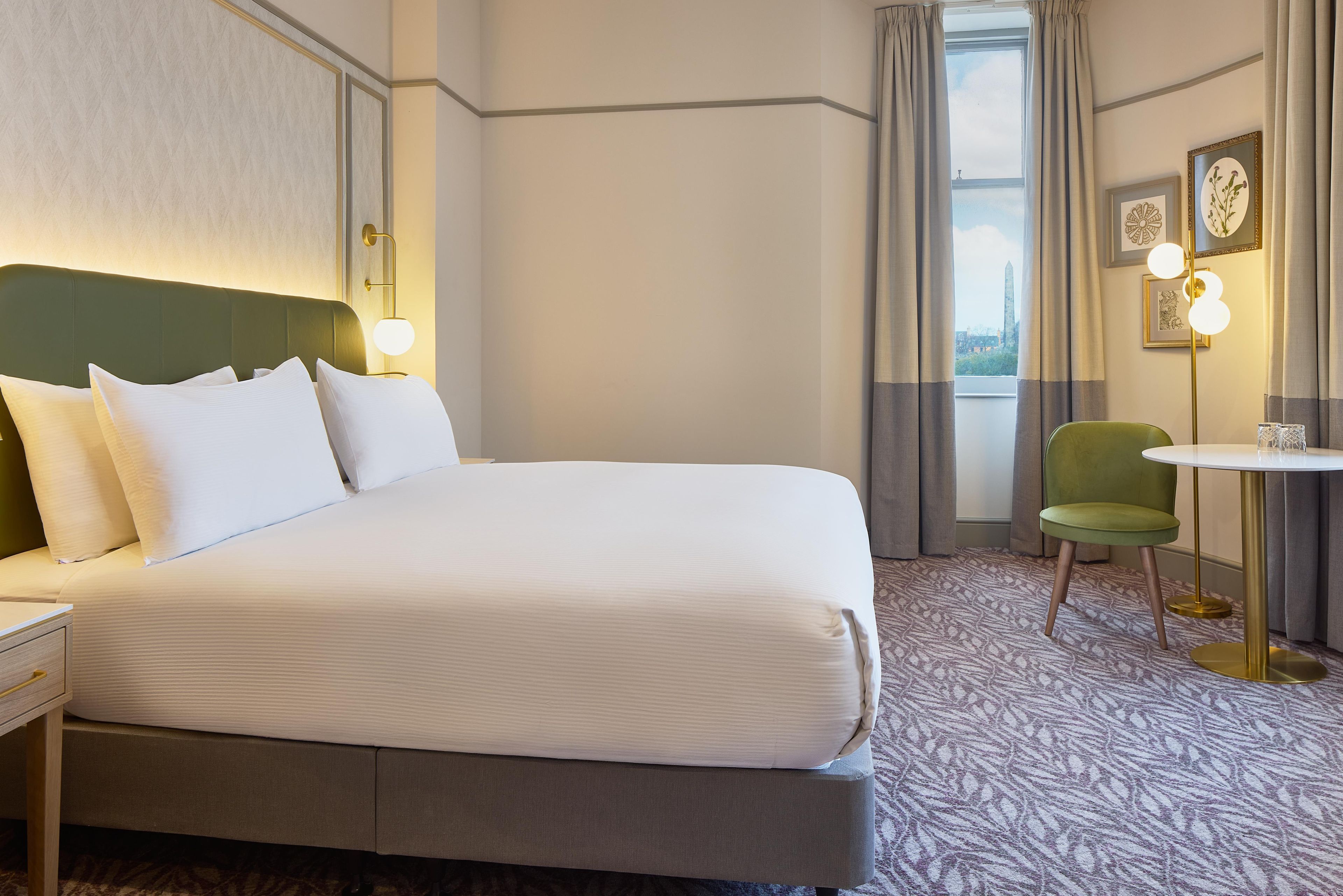 Habitación de hotel con una cama grande, un escritorio, una silla, una mesita y vistas a la ciudad por la ventana.