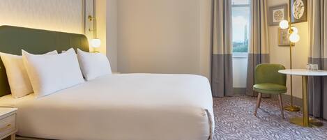 A hotel room with a large bed, a desk, a chair, a small table, and a view of the city through the window.