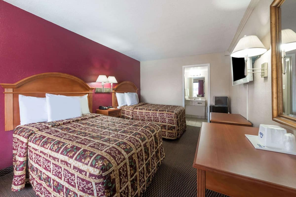 Habitación de hotel con dos camas, un escritorio, un espejo, un televisor y un baño visible a través de una puerta abierta.