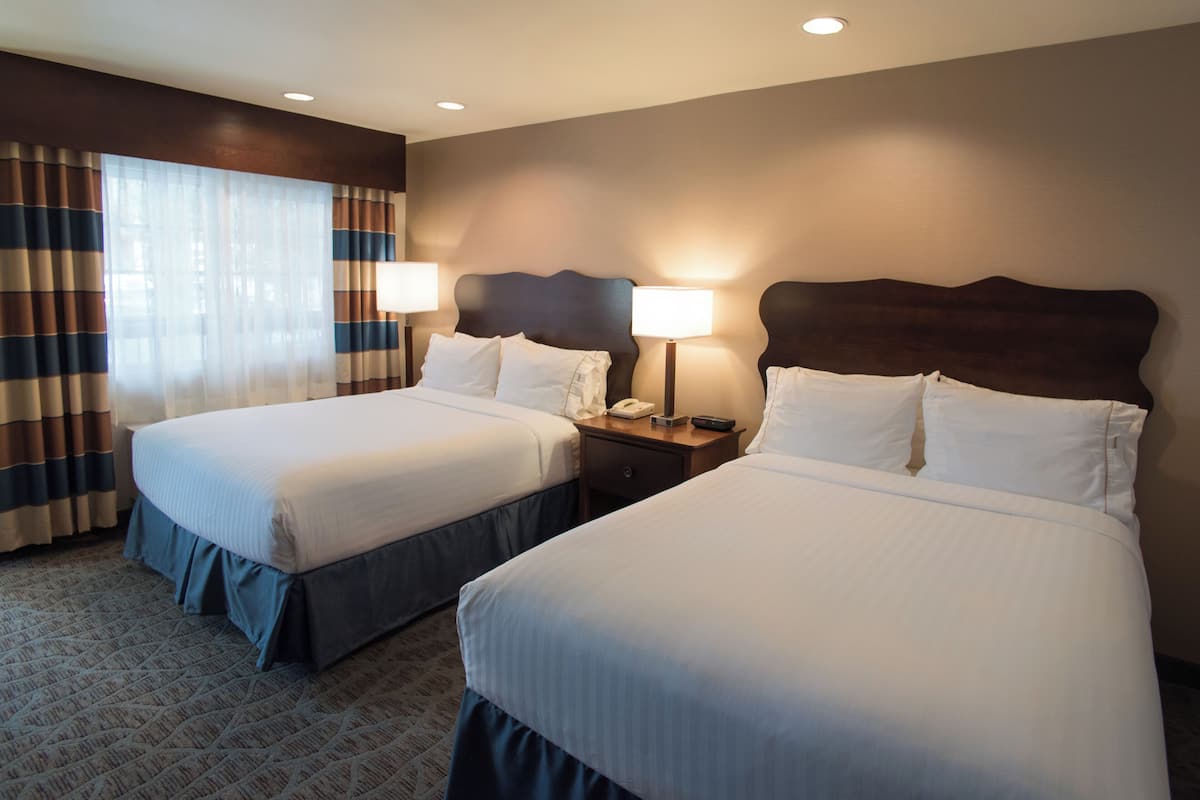 A hotel room with two beds, a nightstand with a lamp, and a window with curtains.