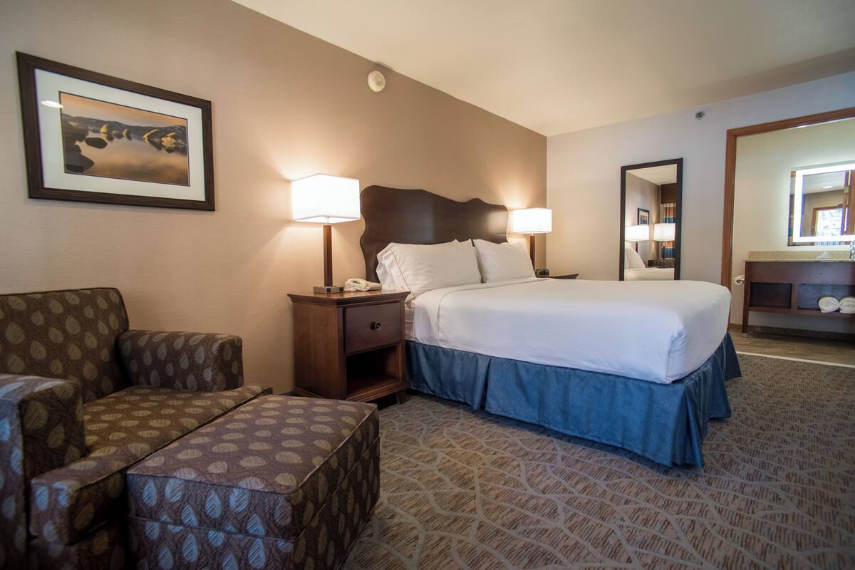 A hotel room with a bed, two bedside lamps, a nightstand, a chair, a small table, and a framed picture on the wall.