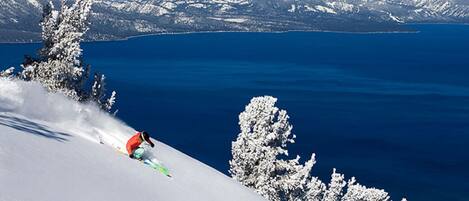 A skier on a snowy slope with a clear view of a lake and mountains in the background.