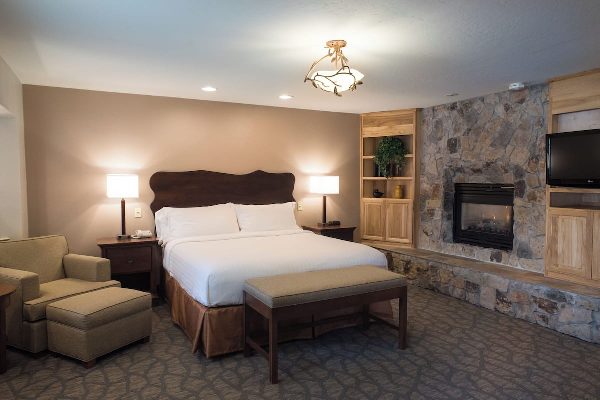 A hotel room with a bed, bedside lamps, a bench, a TV, a fireplace, and a stone wall.
