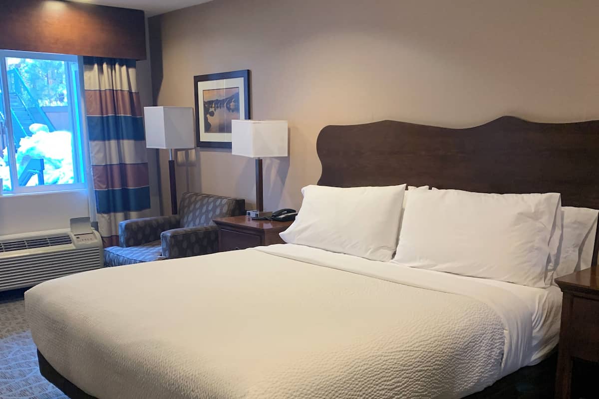A hotel room with a large bed, a nightstand with a lamp, a chair, a window with curtains, and a framed picture on the wall.