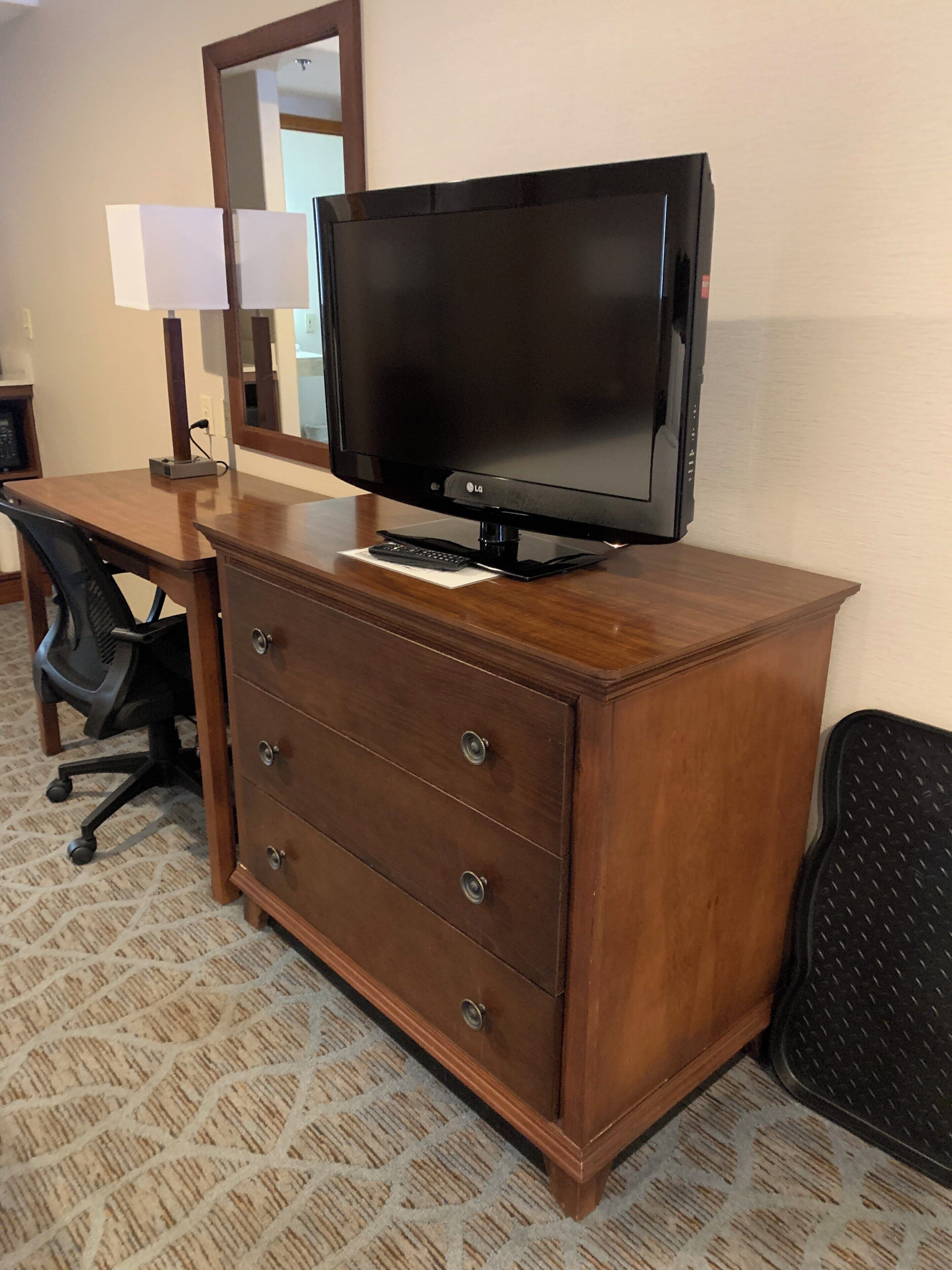 A hotel room with a wooden desk, a flat-screen TV, a mirror, a chair, and a carpeted floor.
