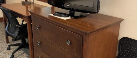 A hotel room with a wooden desk, a flat-screen TV, a mirror, a chair, and a carpeted floor.