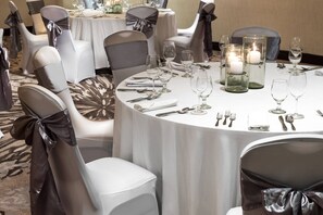 Una mesa redonda para comer con manteles blancos, sillas con cintas grises y negras, y velas en candelabros de vidrio.