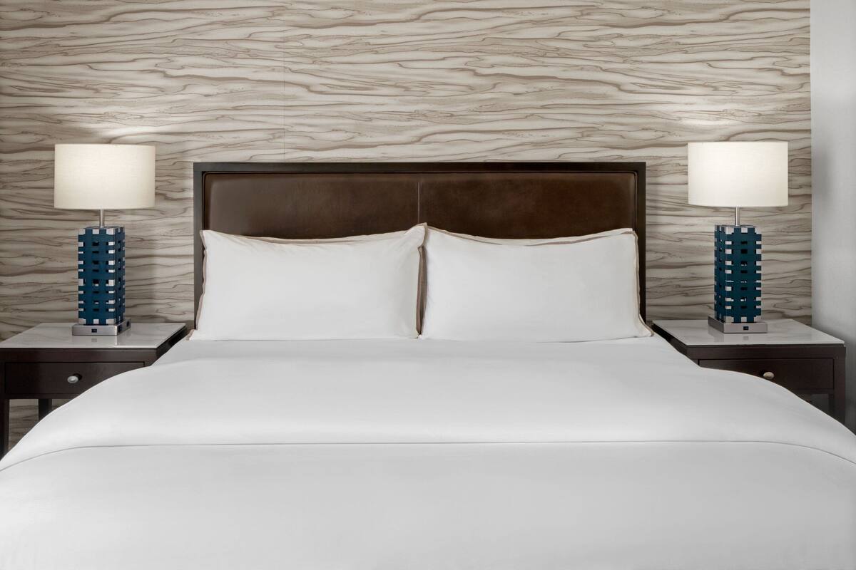Une chambre d’hôtel avec un mur décoré d’un motif imitant le marbre, un grand lit recouvert de draps blancs et deux tables de chevet surmontées de lampes bleues.
