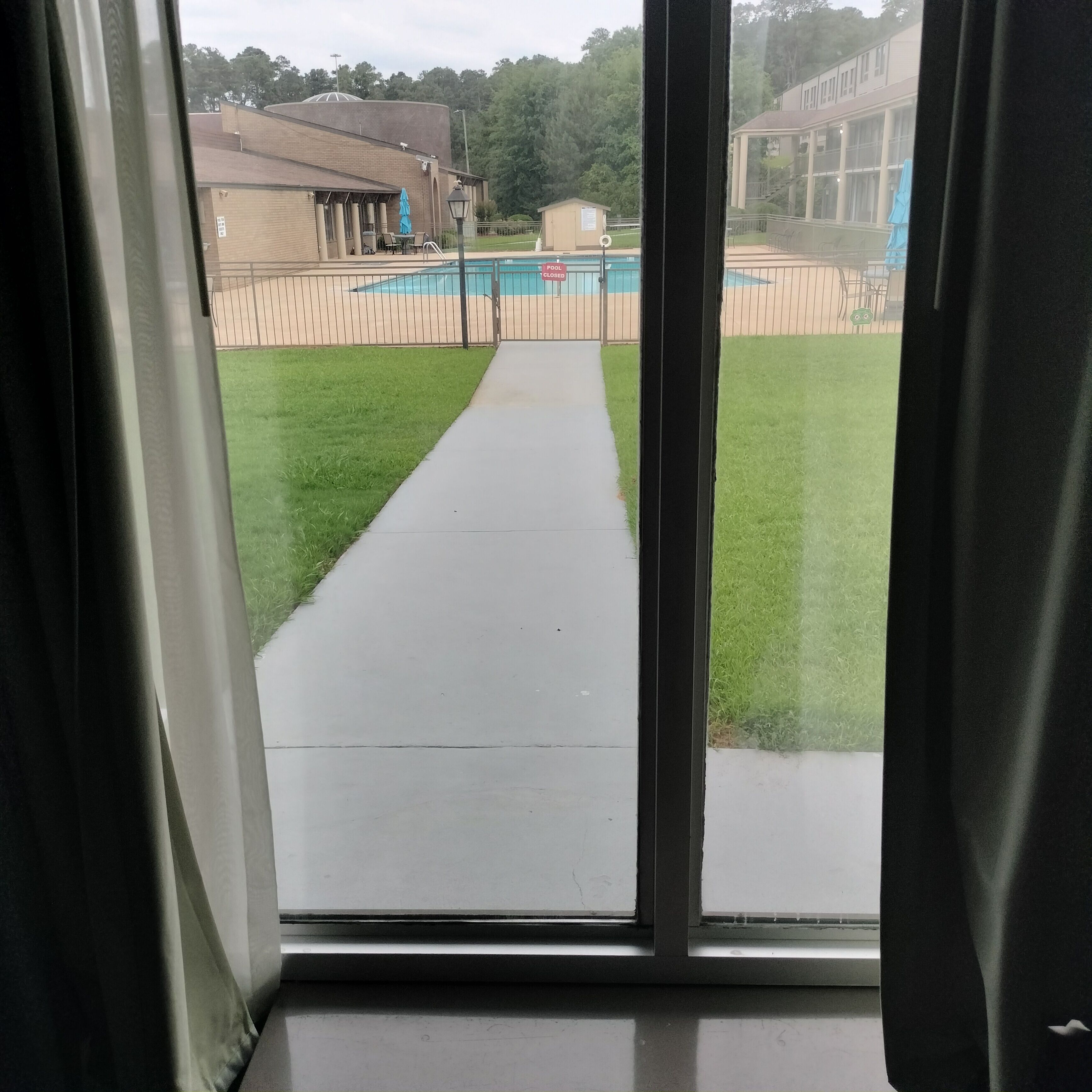 A view through a window of a pool area with a pathway, a building, and a sign indicating the pool is closed.