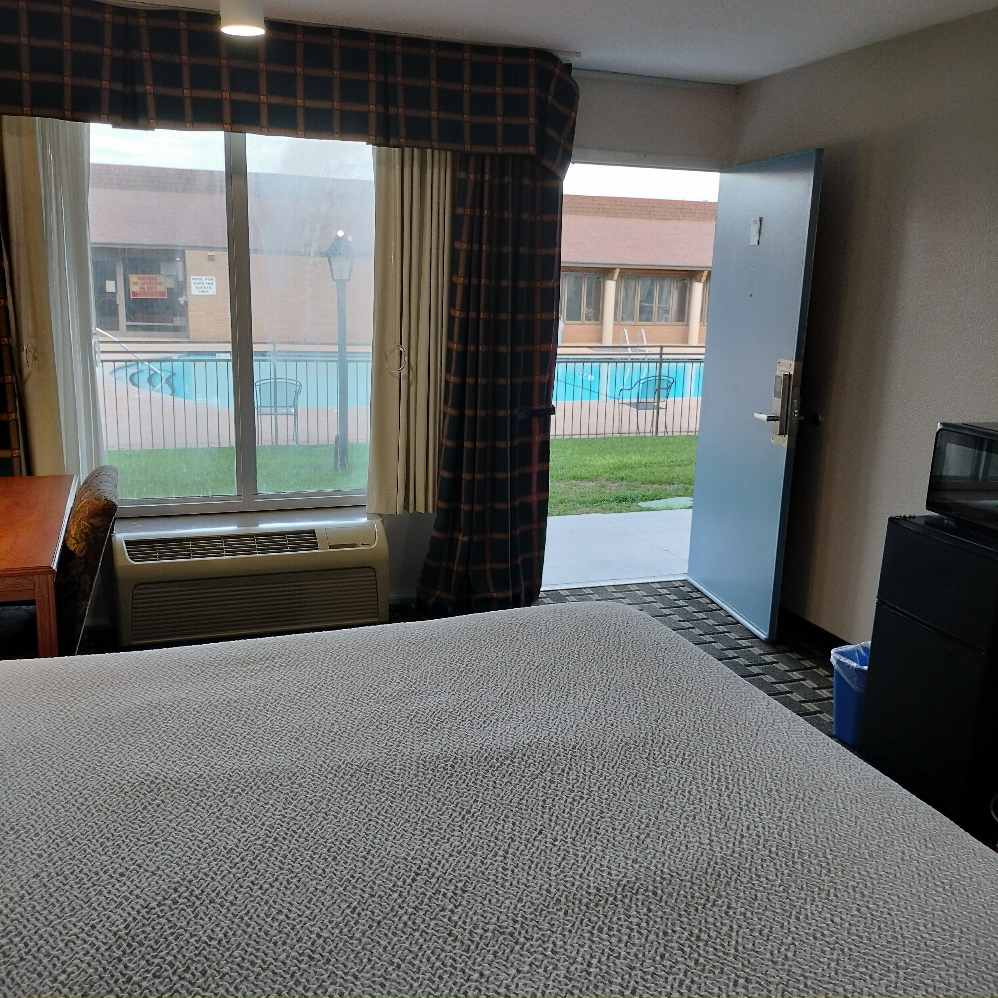 A hotel room with a bed, air conditioning unit, a desk, a chair, a television, and a view of a pool and building outside.
