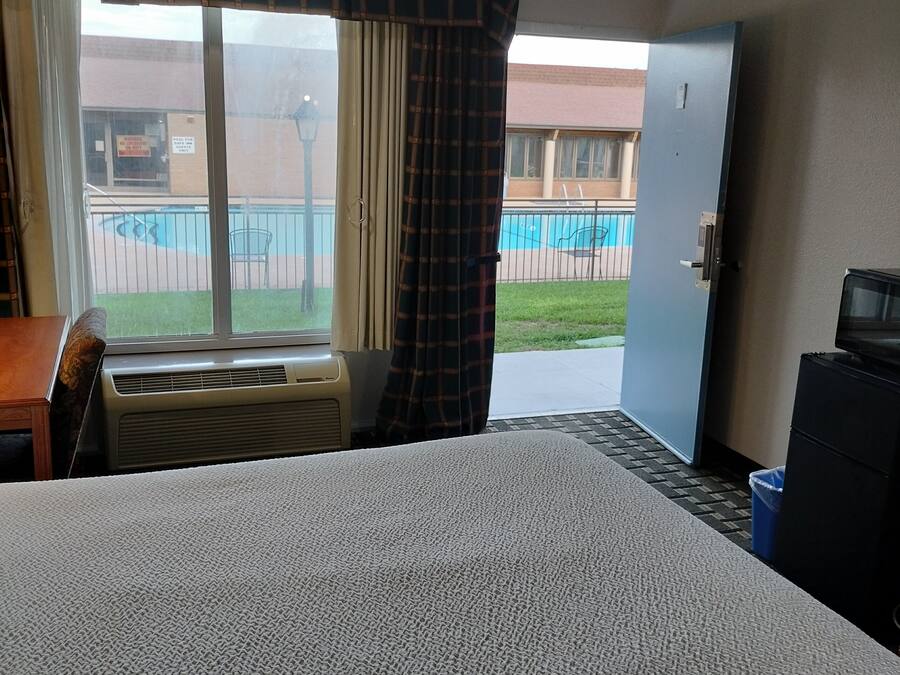 A hotel room with a bed, air conditioning unit, a desk, a chair, a television, and a view of a pool and building outside.