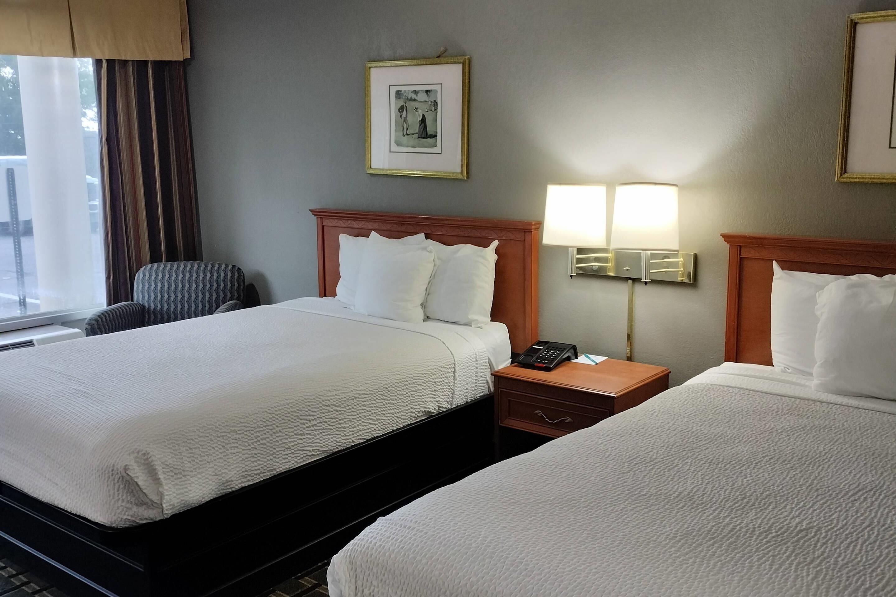 Una habitación de hotel con dos camas, una silla, una mesita de noche con lámpara y un cuadro en la pared.