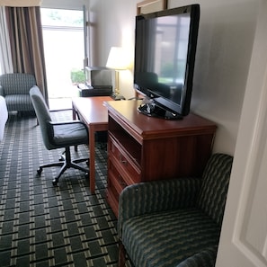 A hotel room with a television, a desk, a chair, a patterned carpet, and a window with curtains. - Clarion Inn Southern Pines - Pinehurst (Southern Pines)