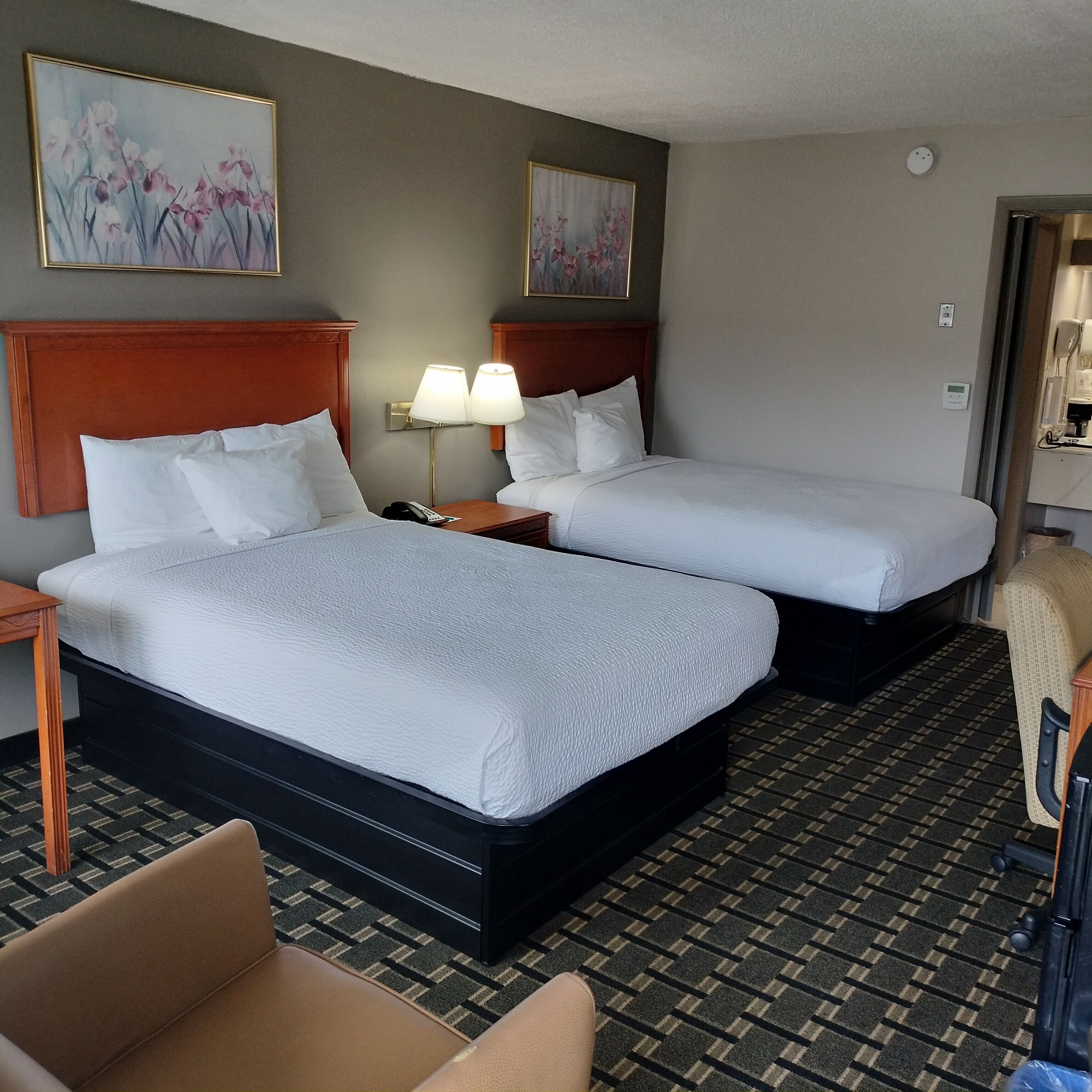 A hotel room with two beds, a nightstand with a lamp, a chair, and artwork on the walls.