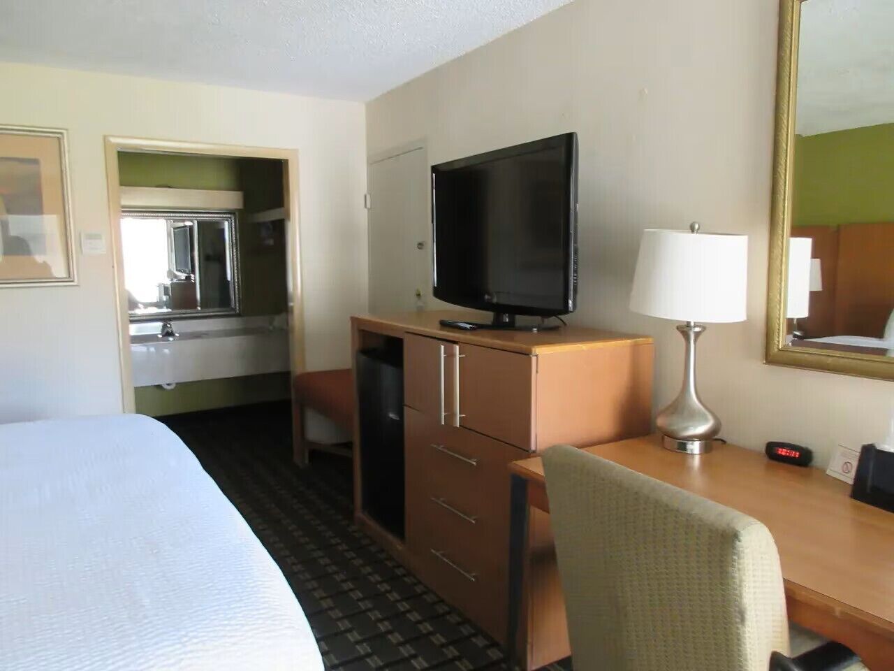 A hotel room with a bed, a desk with a television, a chair, a desk lamp, and a mirror.