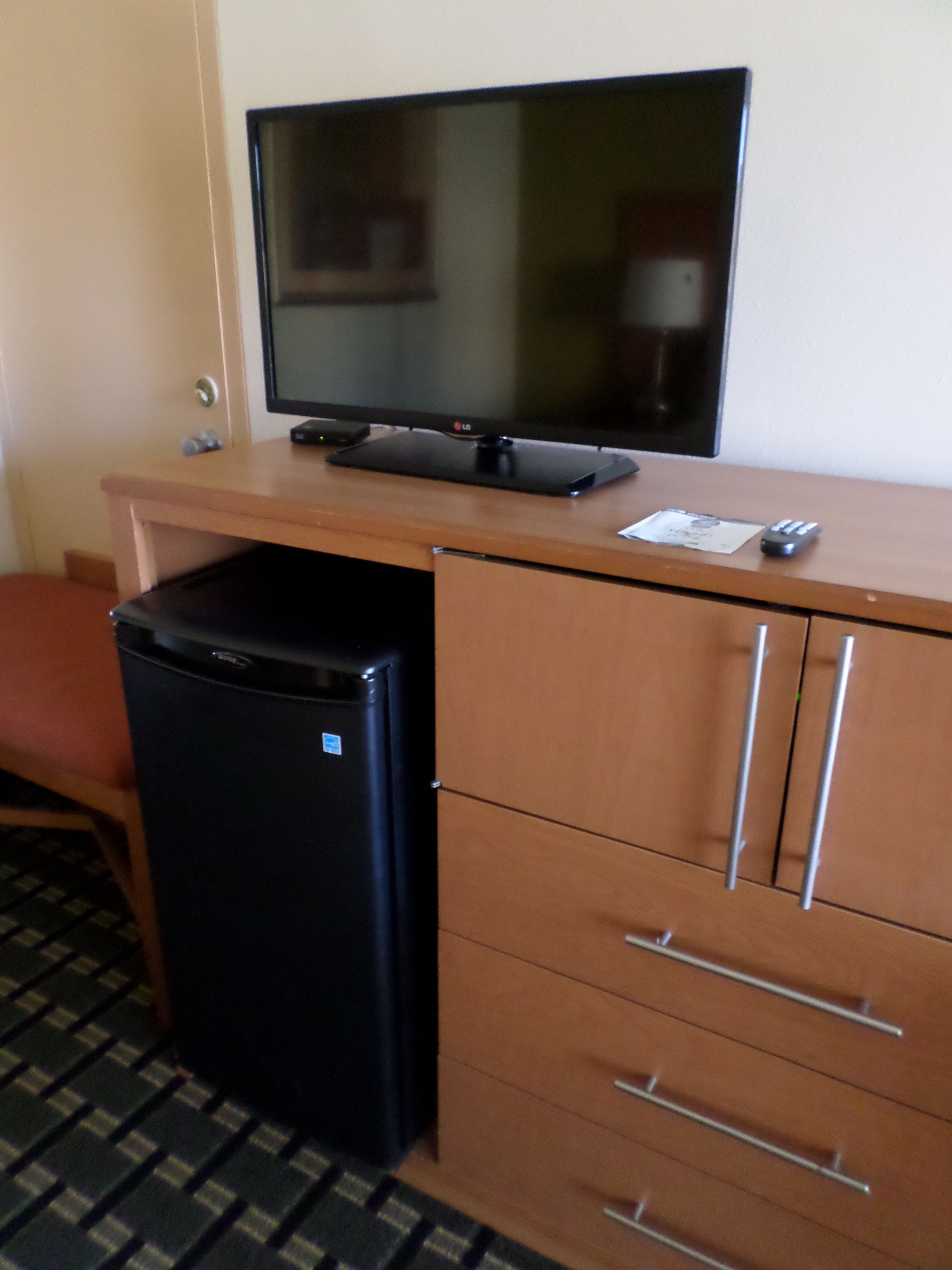 A compact kitchenette with a refrigerator, a small black appliance, a flat-screen TV on a wooden desk, and a chair.