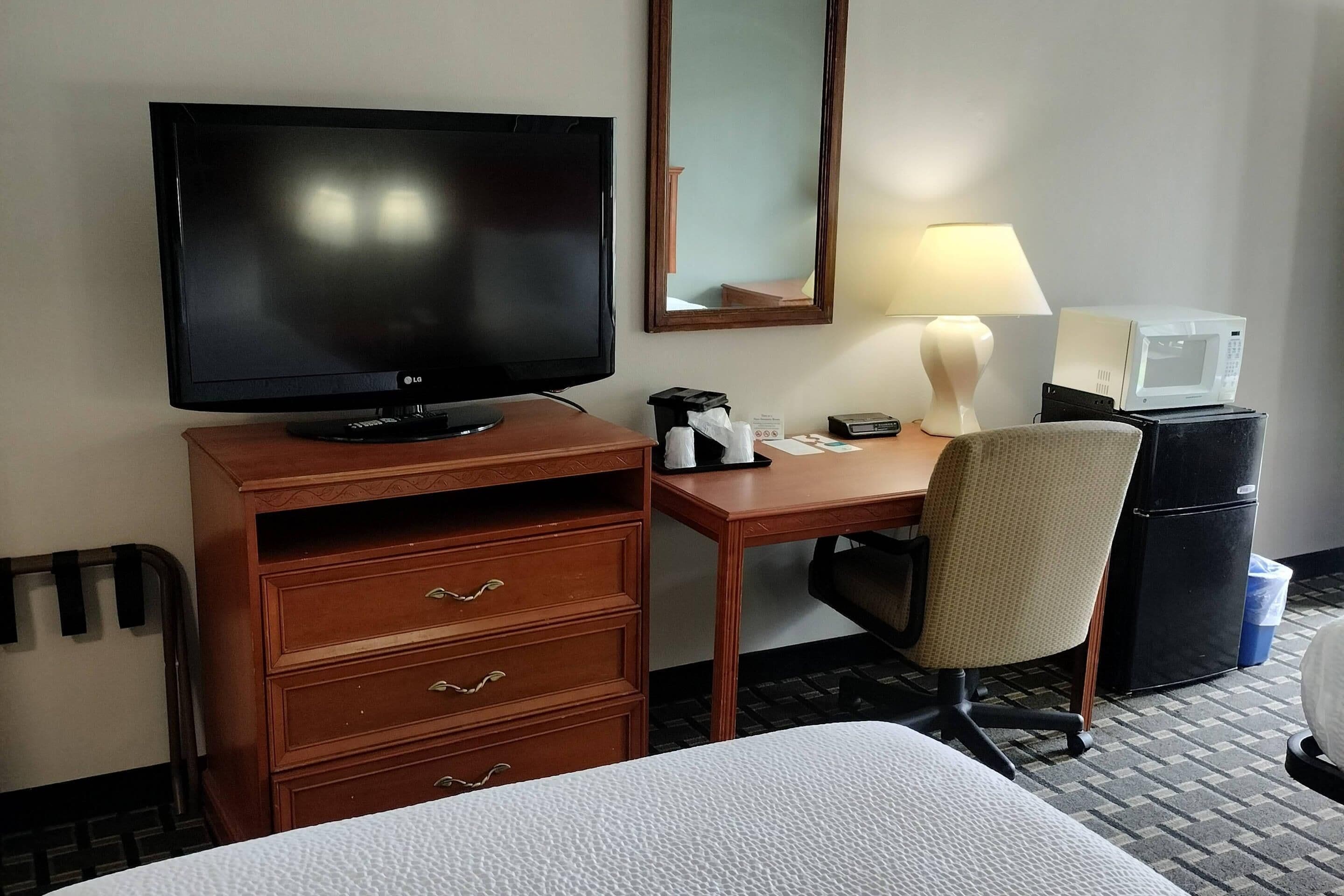 A hotel room with a bed, a desk with a television, a chair, a microwave, and a refrigerator.