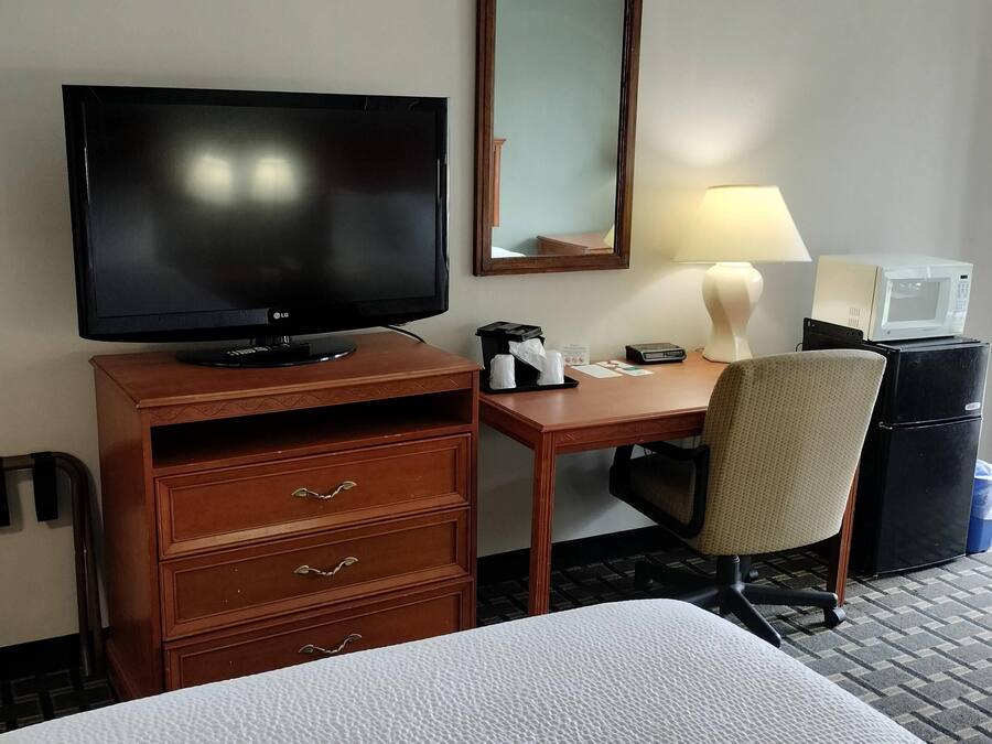 A hotel room with a bed, a desk with a television, a chair, a microwave, and a refrigerator.