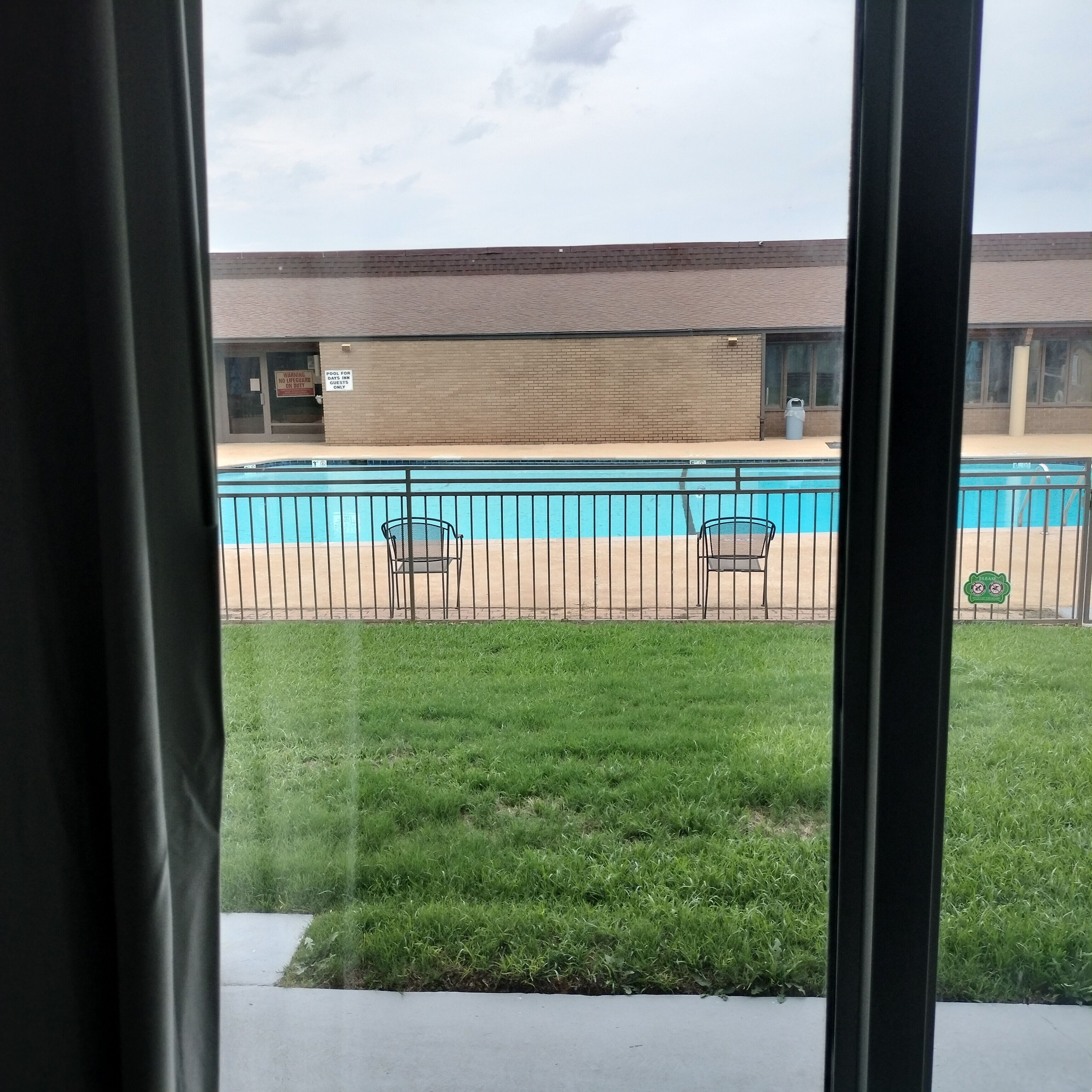 A view of a swimming pool with a fence, a grassy area, and a building with a brick facade.