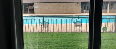 A view of a swimming pool with a fence, a grassy area, and a building with a brick facade.