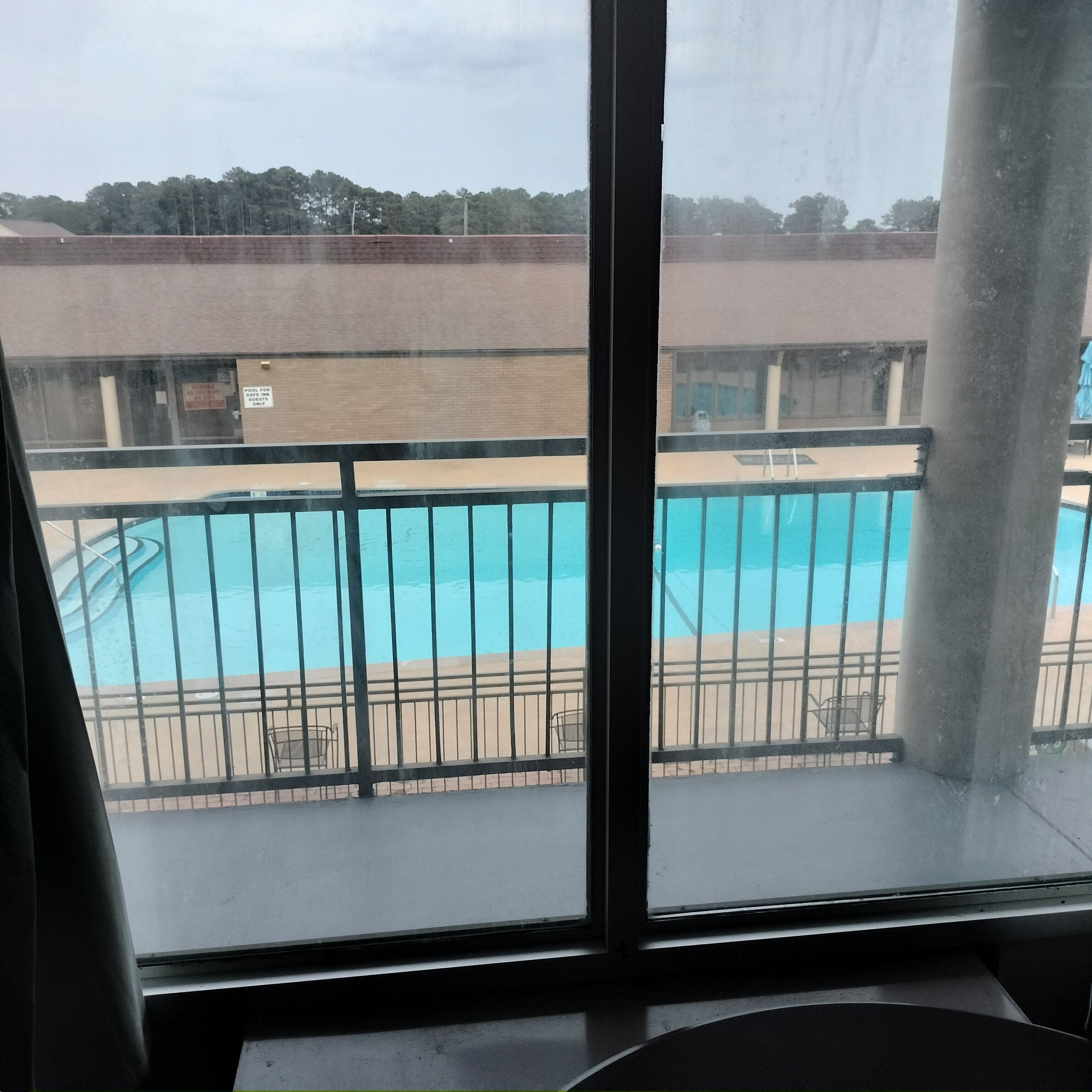 A view of a swimming pool from a window with a metal railing, a brick wall, and a building in the background.
