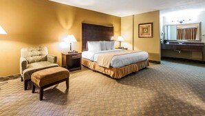 Une chambre d’hôtel avec un lit, une chaise, une table de chevet, une lampe, un cadre photo et un miroir dans la salle de bain.